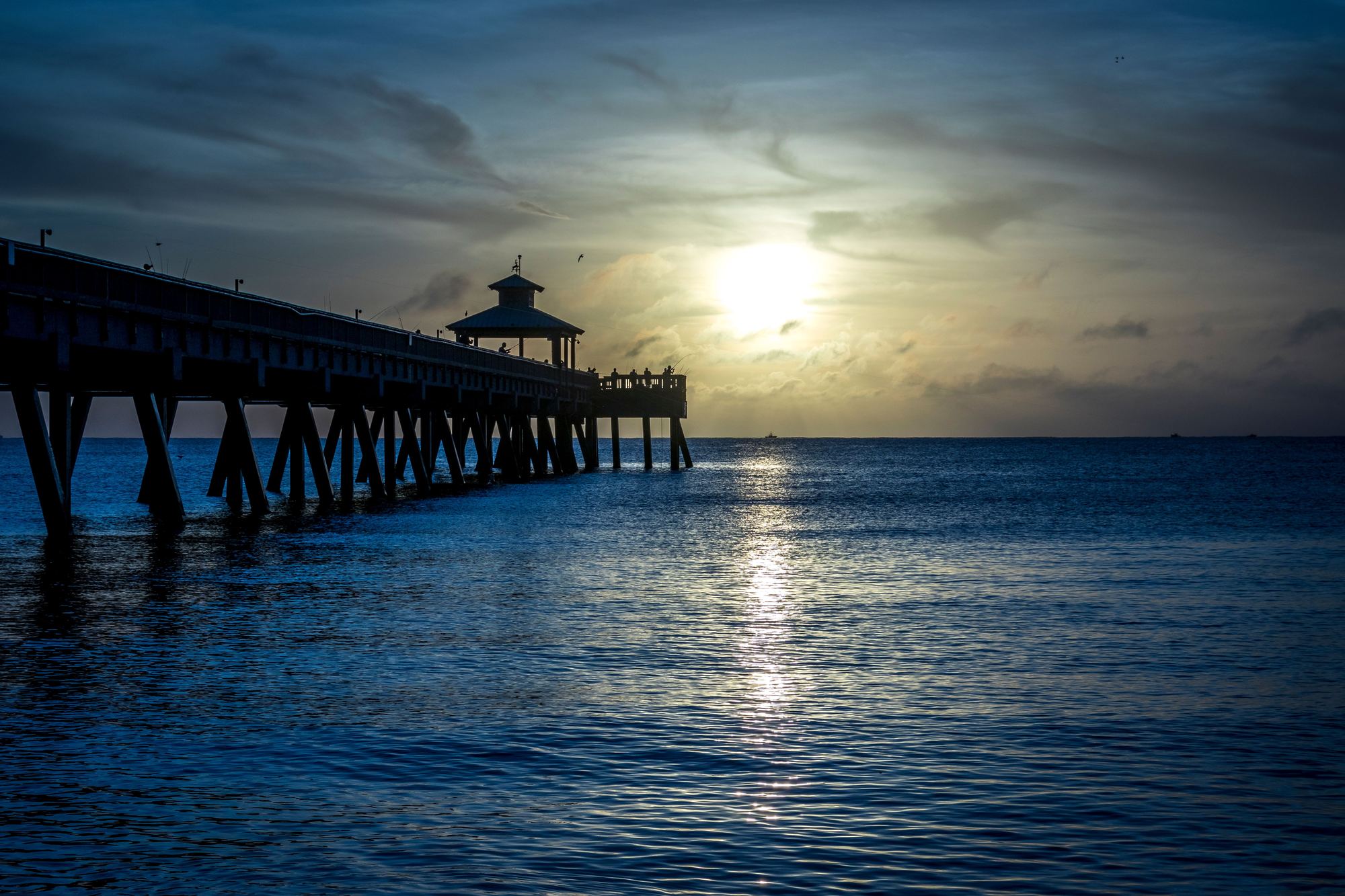 The Deerfield Beach fishing pier silhouetted over the Atlantic, with a low sun and its reflection on the water.