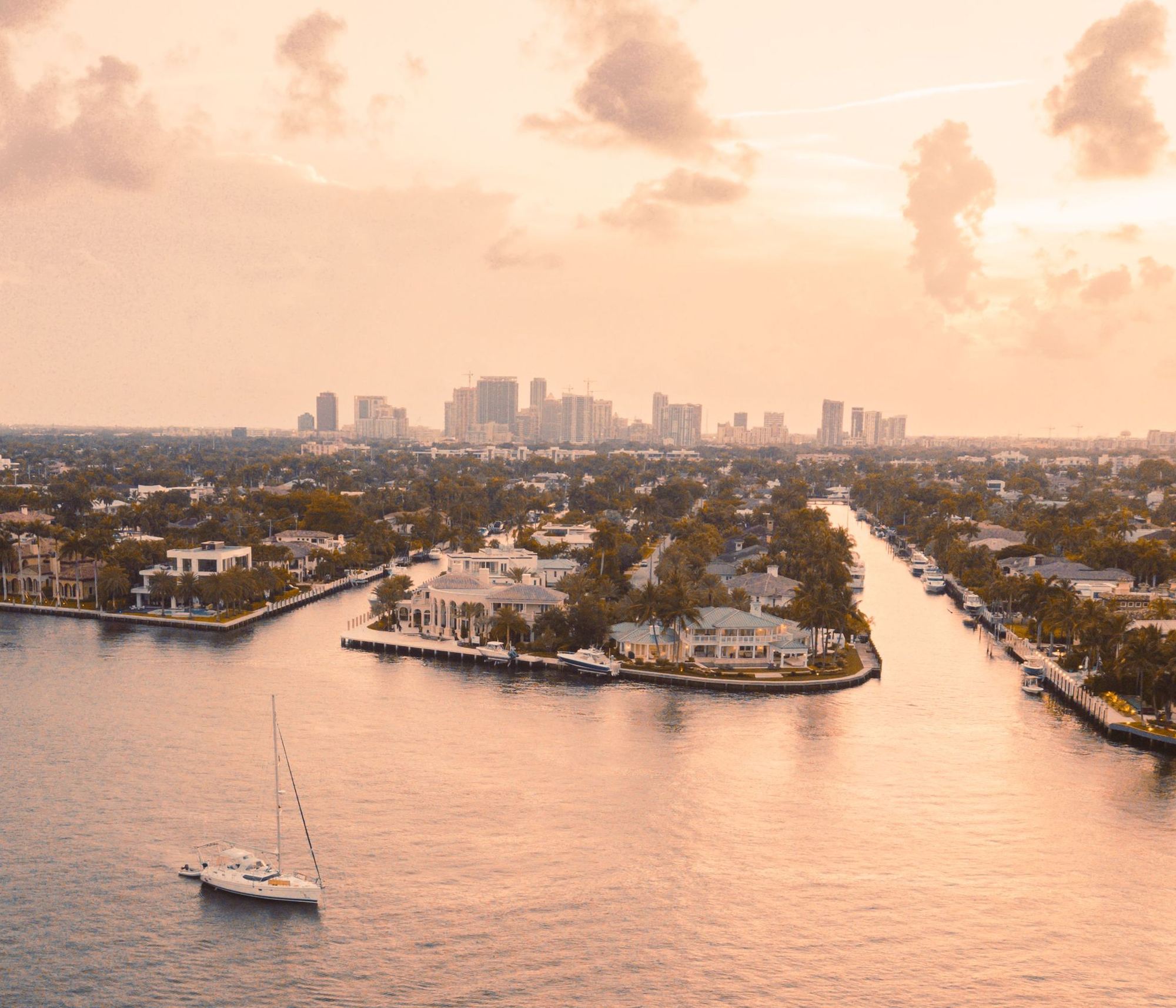 Aerial view of Fort Lauderdale at sunset, with the downtown skyline beyond canal-front homes and a sailboat on the water.